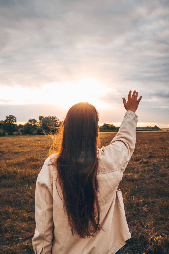 Mulher de costas com a mão erguida em um campo ao pôr do sol